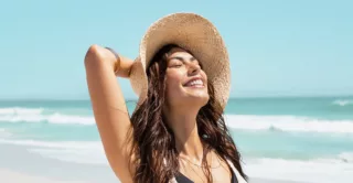 A smiling woman with glowing skin at the beach wearing a sun hat
