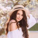 A young woman with glowing skin in a white dress and straw hat standing in front of flowers, smiling in the sun, showing the concept of aesthetic treatments like Botox, Juvederm, and more