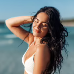 A young woman with dark brown hair and glowing skin closes her eyes and smiles in the sun on the beach