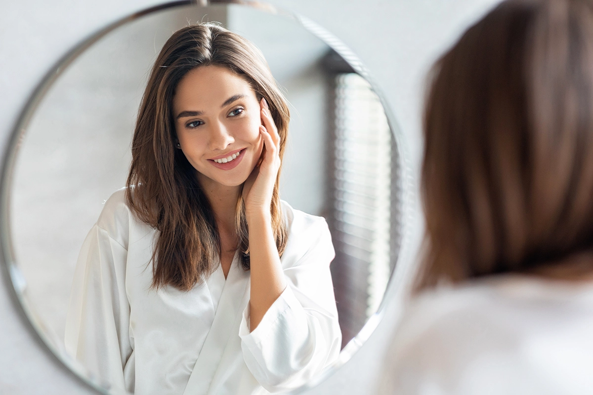A woman smiles while touching her face in the mirror, showing the concept of Sofwave skin rejuvenation