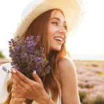 A young woman smiling in the sun, wearing a sun hat and holding lavender flowers