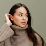 A woman tucks her hair behind her ear, wearing a light brown knit sweater