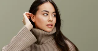 A woman tucks her hair behind her ear, wearing a light brown knit sweater