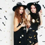 Two smiling woman standing side by side with drinks in witch costumes, against a white background with bat confetti, showing the concept of halloween