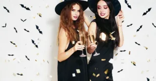 Two smiling woman standing side by side with drinks in witch costumes, against a white background with bat confetti, showing the concept of halloween