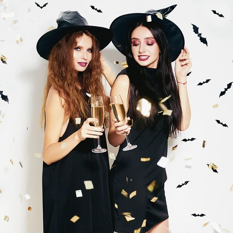 Two smiling woman standing side by side with drinks in witch costumes, against a white background with bat confetti, showing the concept of halloween