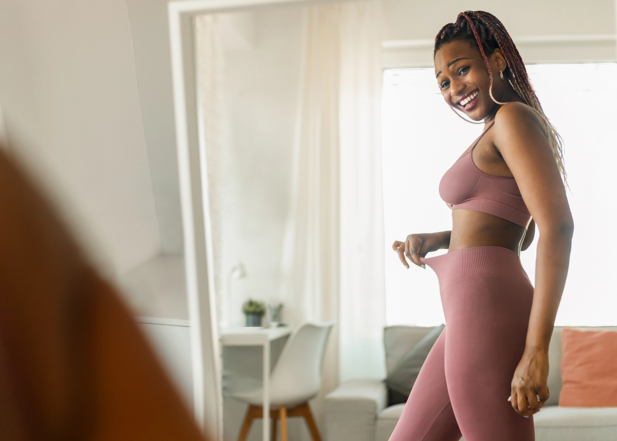 A smiling woman looking in the mirror with loose leggings, showing the concept of weight loss injections