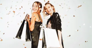 Two smiling women in black dresses holding shopping bags for Black Friday