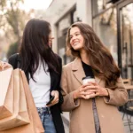 Two smiling friends carrying shopping bags and coffee, with fall trees in the background