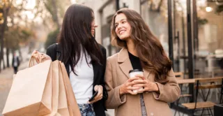 Two smiling friends carrying shopping bags and coffee, with fall trees in the background