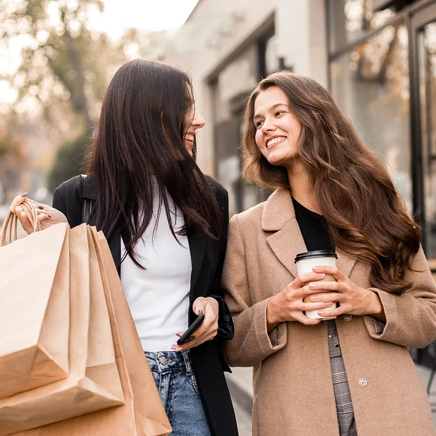 Two smiling friends carrying shopping bags and coffee, with fall trees in the background