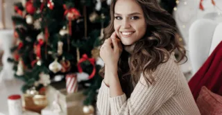 A beautiful women wearing a turtleneck sweater, sitting in front of a Christmas tree with gifts.