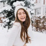 A smiling young woman with long brown hair wearing white winter clothes in the snow