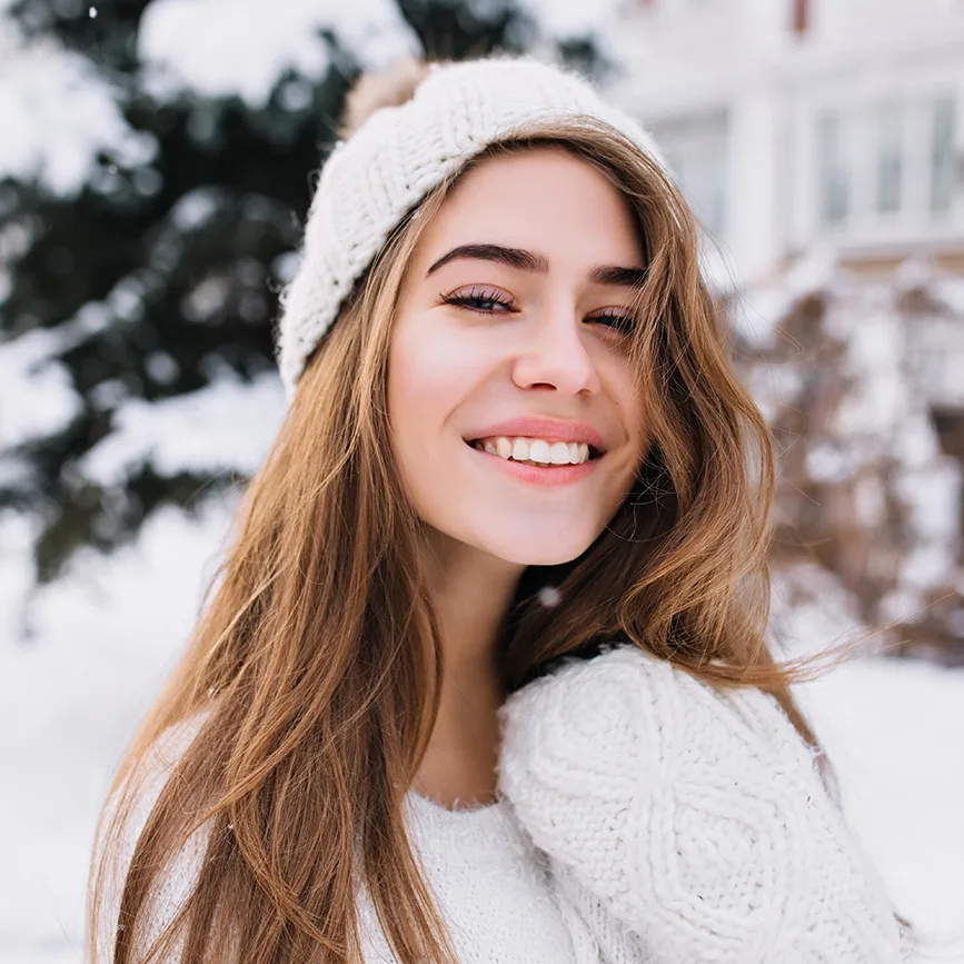 A close up of a smiling young woman with long brown hair, wearing white winter clothes in the snow