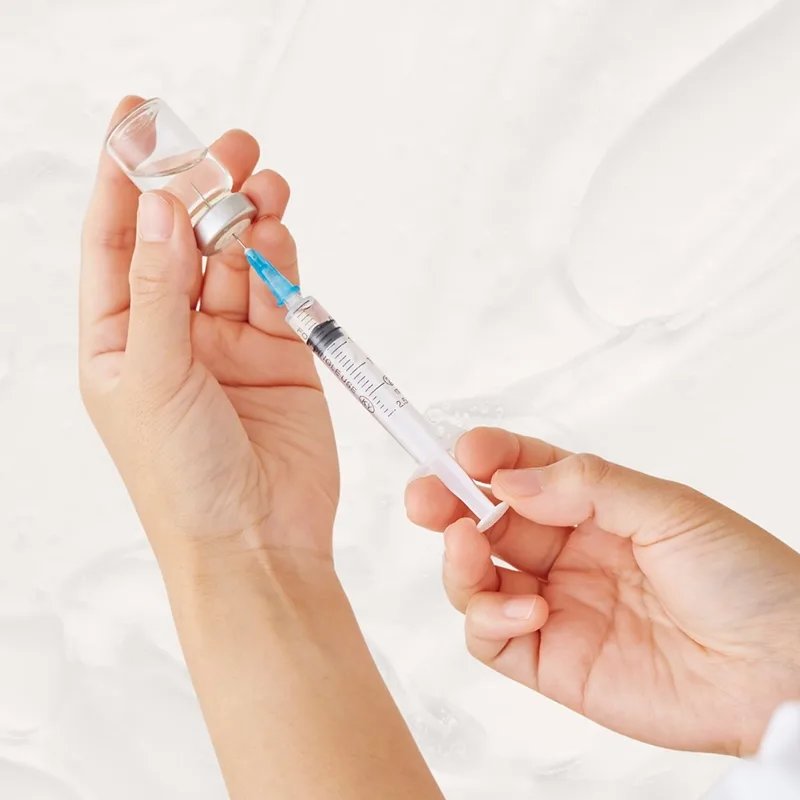 Two hands preparing a peptide or enzyme therapy injection over a cream colored background