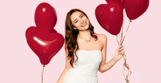 A young woman in a white dress holding red heart-shaped balloons against a pink background, showing the concept of Valentine's Day