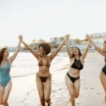 Four beautiful women in swimsuits smiling and laughing at a beach during sunset.