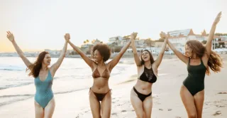Four beautiful women in swimsuits smiling and laughing at a beach during sunset.