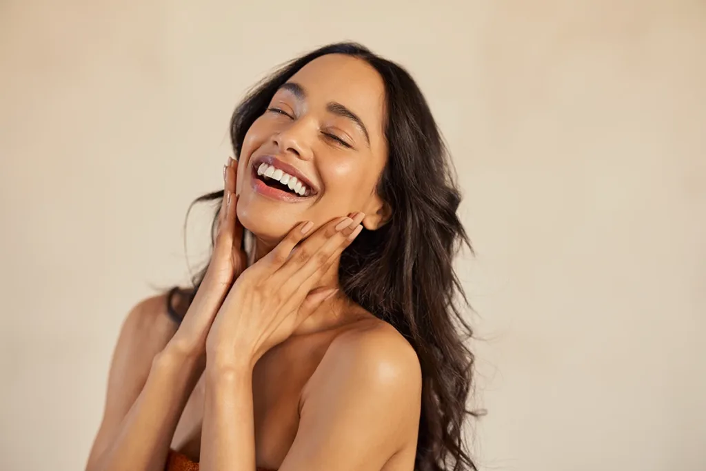A beautiful woman with long dark hair, and perfect, healthy skin, touching face smiling; anti-aging concept 