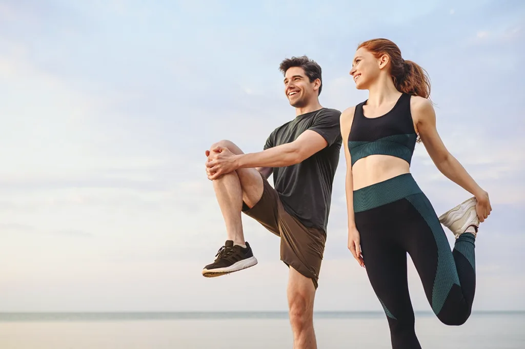 A man and a woman wearing athletic clothing stretch on a beach, showing the concept of exercise and energy