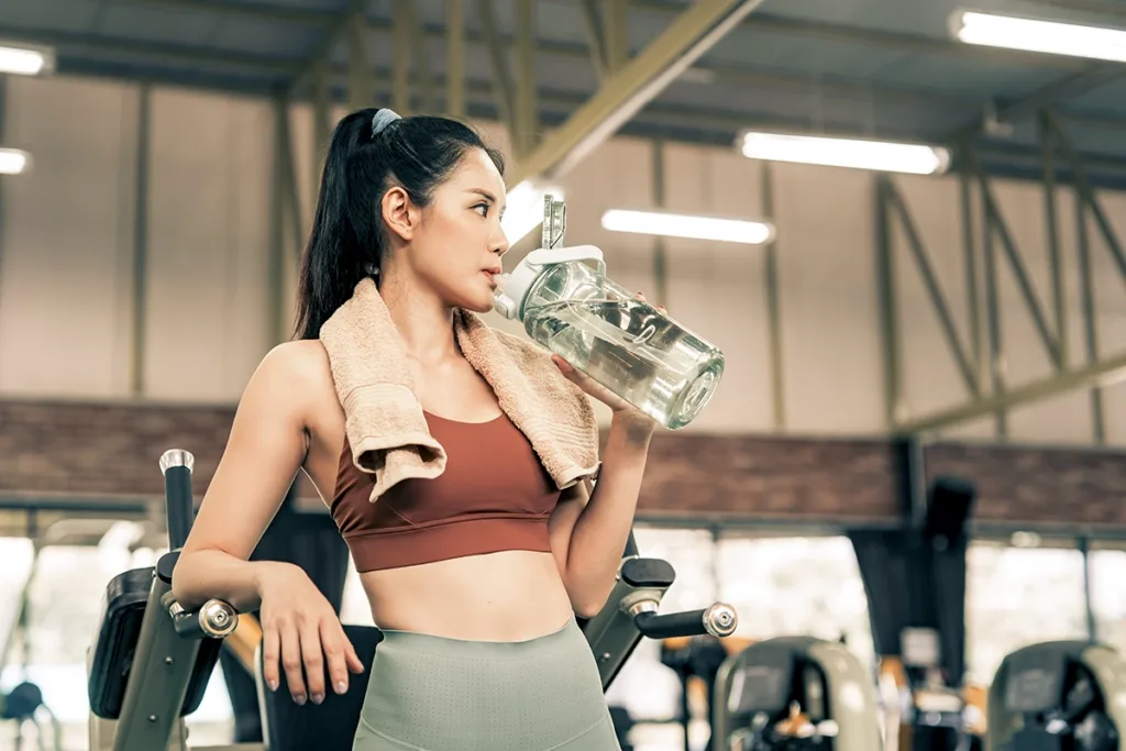 An athletic woman in a gym drinks from a clear water bottle, showing the concept of hydration