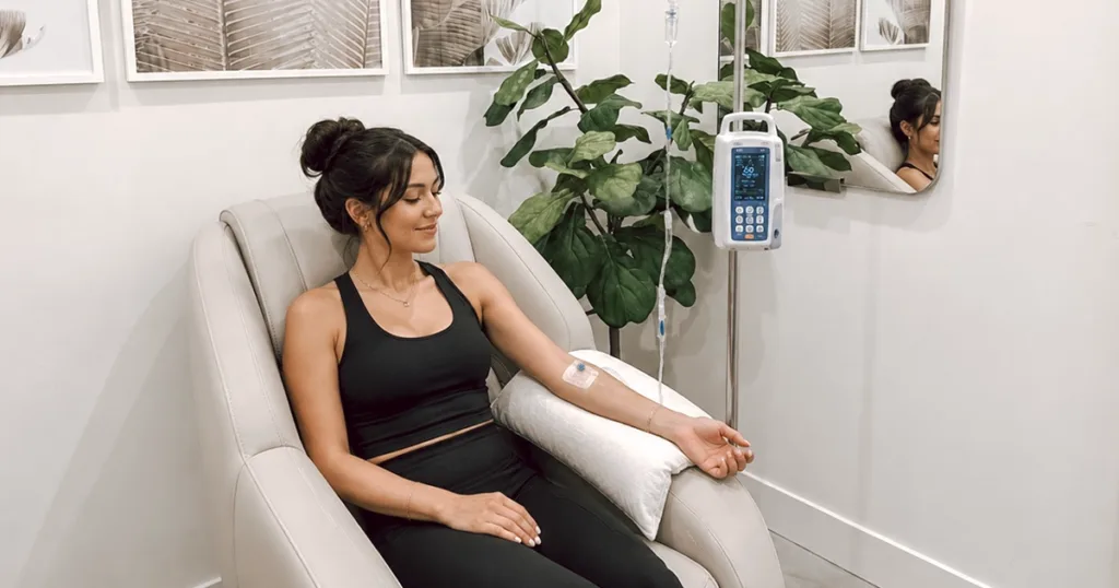 A smiling woman in a med spa clinic receives IV therapy for hydration and wellness, with a ficus plant in the background