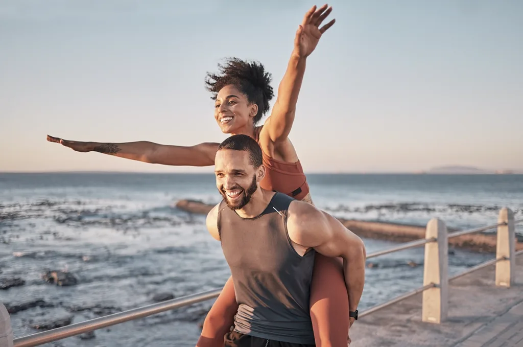 A smiling man holds a woman on his back, who has her arms outstretched, with an ocean in the background, showing the concept of wellness