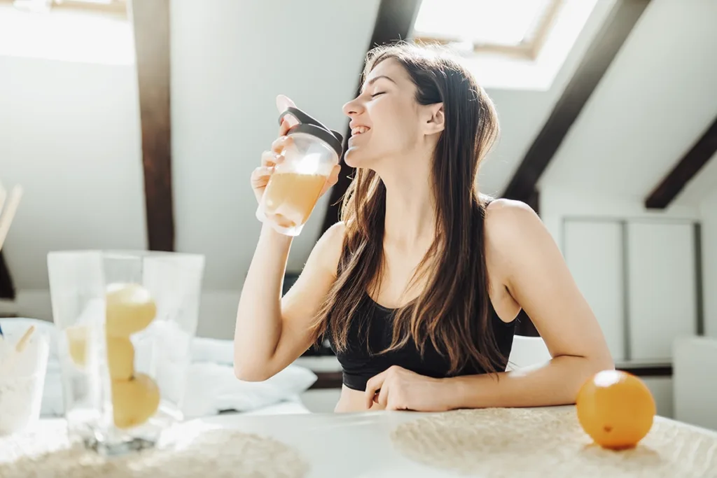 A young woman wearing athletic clothes smiles while drinking orange juice at a table, showing the concept of immunity and wellness