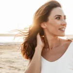 A smiling woman with brown hair in a white top stands in the sun at the beach, showing the concept of summer