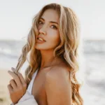 A woman with blonde hair stands in the sunlight at a beach, twirling her hair, showing the concept of summer