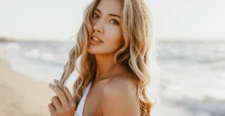 A woman with blonde hair stands in the sunlight at a beach, twirling her hair, showing the concept of summer
