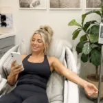 A smiling young woman with her hair up checks phone while receiving IV therapy treatment at a med spa