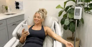 A smiling young woman with her hair up checks phone while receiving IV therapy treatment at Artemedica med spa in Santa Rosa, California.