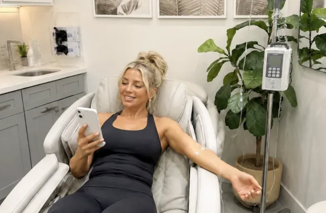 A smiling young woman with her hair up checks phone while receiving IV therapy treatment at a med spa
