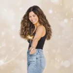 A smiling woman with long curly hair, wearing a black tank top and blue jeans while holding a sparkler, against a beige bokeh background