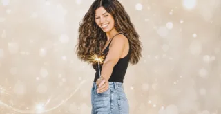 A smiling woman with long curly hair, wearing a black tank top and blue jeans while holding a sparkler, against a beige bokeh background