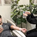 A young man checks his phone while receiving IV therapy treatment at a med spa clinic, with a fiddle leaf fig tree in the background