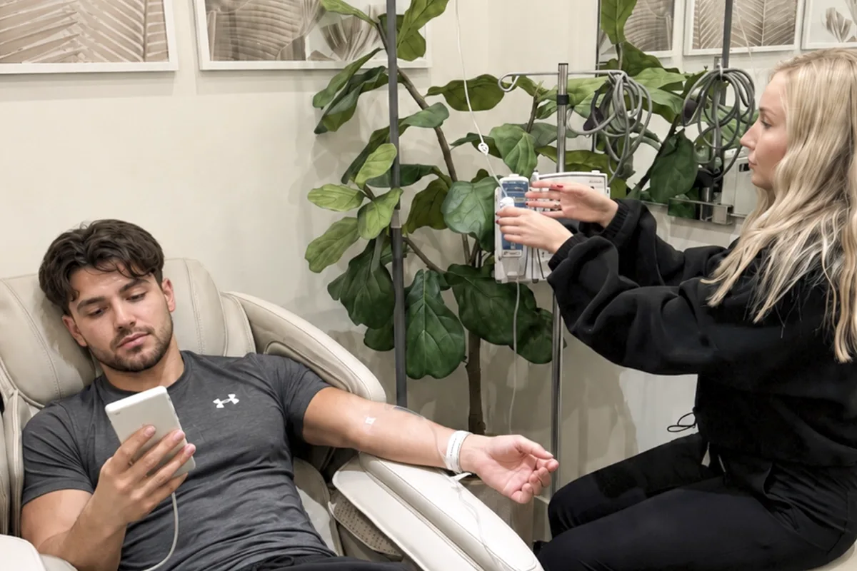 A young man checks his phone while receiving IV therapy treatment at a med spa clinic, with a fiddle leaf fig tree in the background