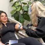 A young woman receives IV therapy treatment from a med spa clinic in a lounge chair, with a fiddle leaf fig tree in the background