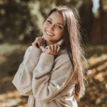 A smiling woman with clear skin wearing a beige knit sweater, with autumn leaves and trees in the background, showing the concept of med spa treatments