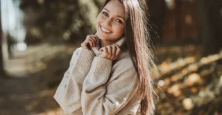 A smiling woman with clear skin wearing a beige knit sweater, with autumn leaves and trees in the background, showing the concept of med spa treatments