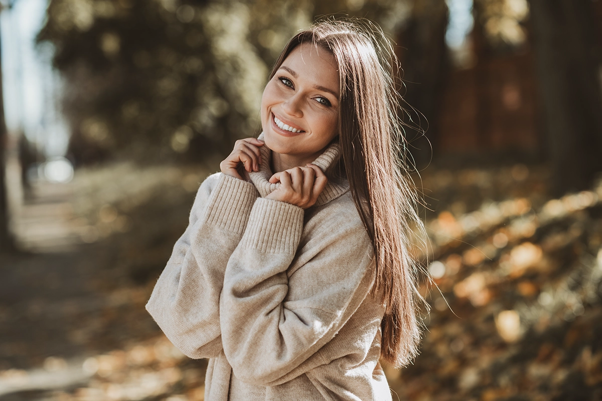 A smiling woman with clear skin wearing a beige knit sweater, with autumn leaves and trees in the background, showing the concept of med spa treatments
